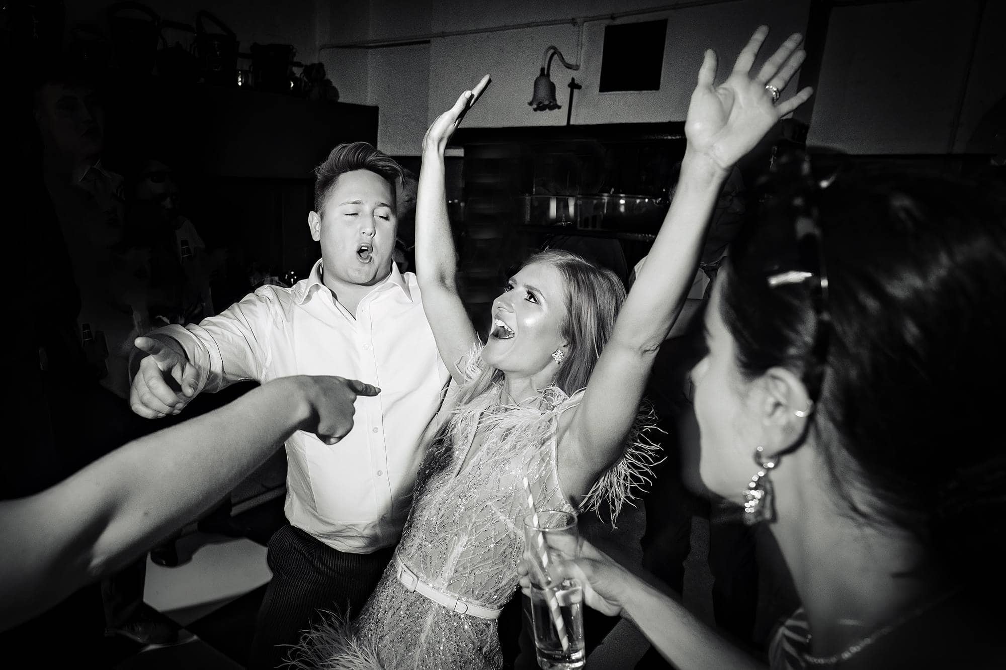Black & white photo of a bride at Belvoir Castle dancing at the evening wedding reception with her hands in the air whilsst a man in a white shirt is behind her and a guest's arm is pointing towards her from the left