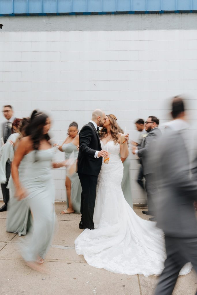 Editorial style photo of bride & groom kissing whilst holding their bubble teas whilst their guests run around them randomly whilst blurry