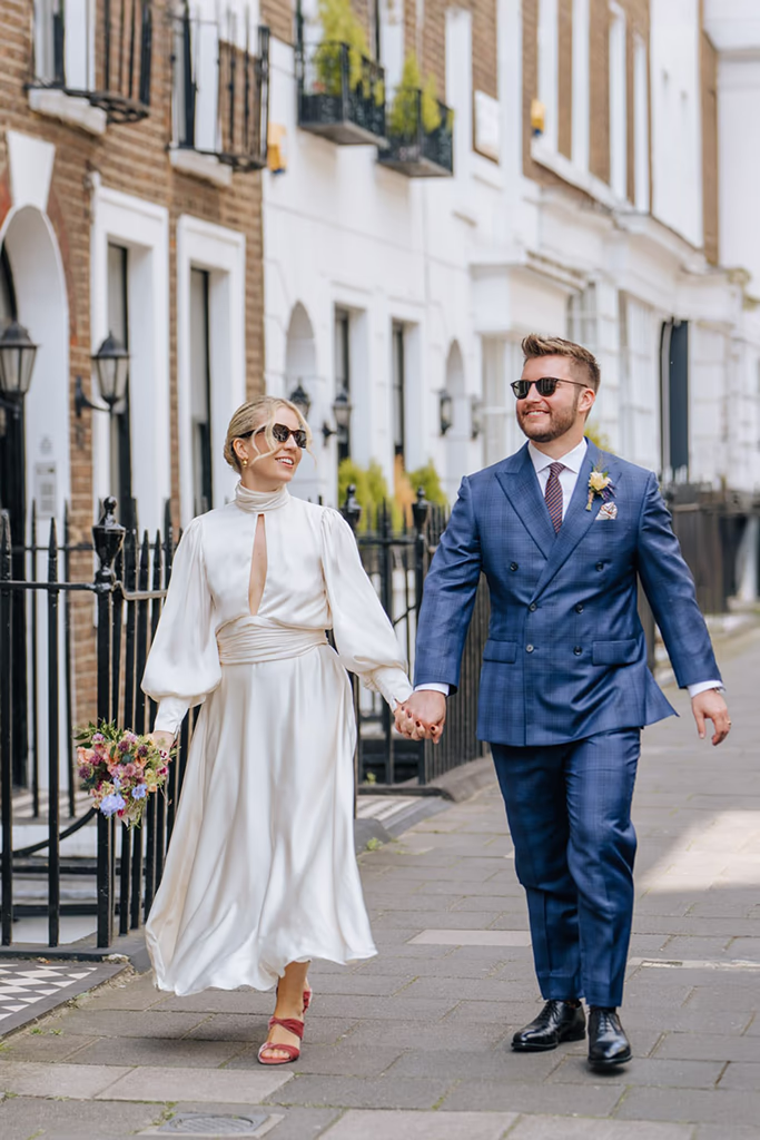 Bride in modern white dress and groom in blue suit wearing sunglasses walking down a city street holding hands. Editorial style wedding portrait by Nottingham wedding photographer Martin Cheung
