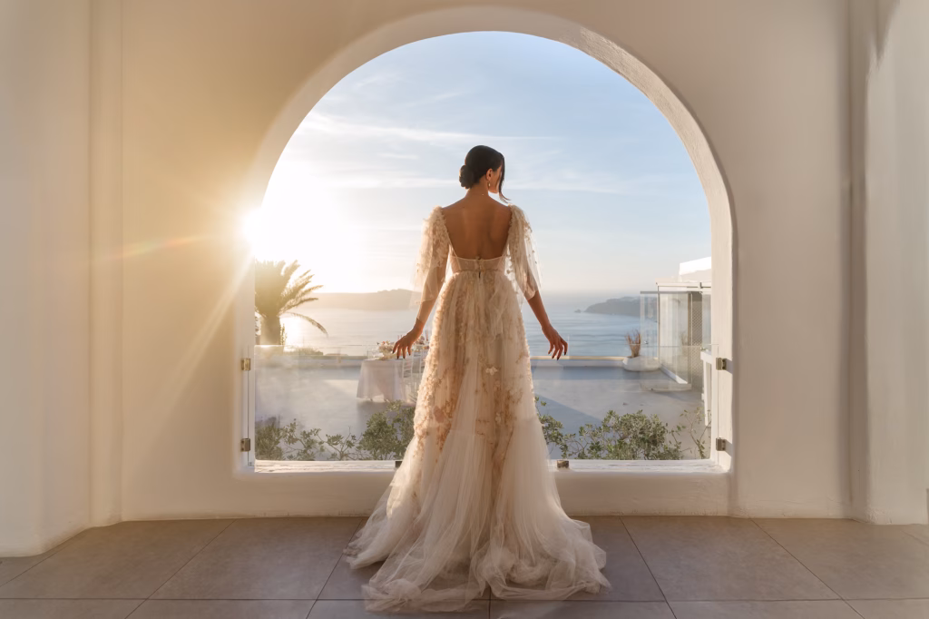 A bride with an elegant wedding dress stands in front of an arch with the golden sunlight peeking through the top left of the archway
