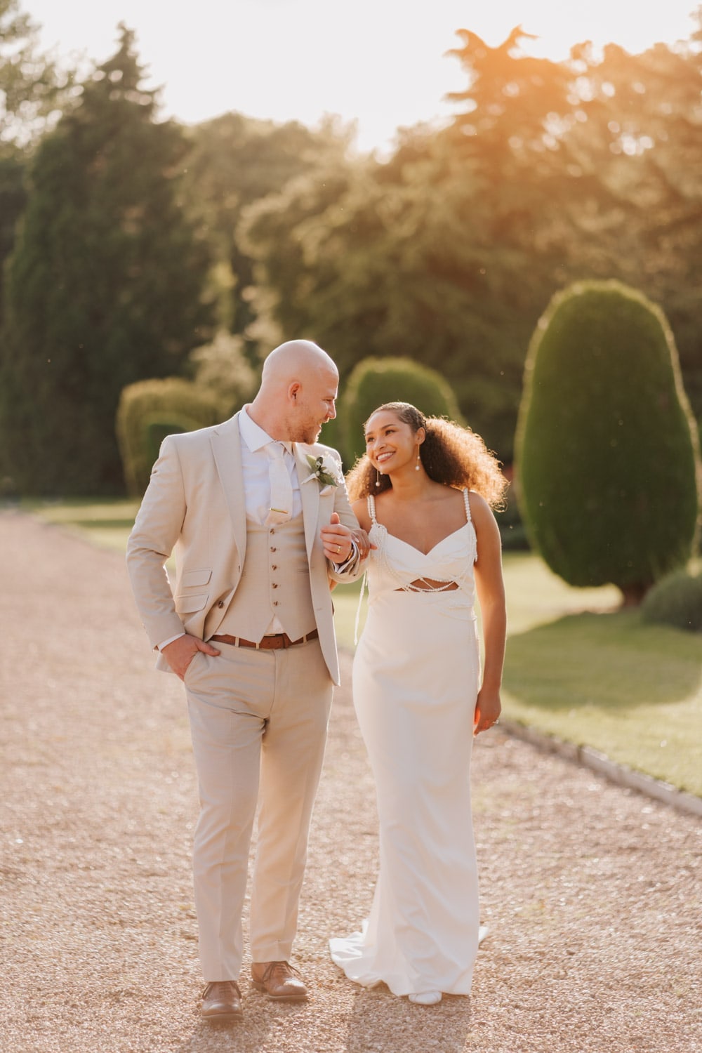 A bride and groom walk outside Prestwold Hall during golden hour, smiling at each other. Captured by Nottingham wedding photographer Martin Cheung —the groom in a tan suit, the bride in a white dress.