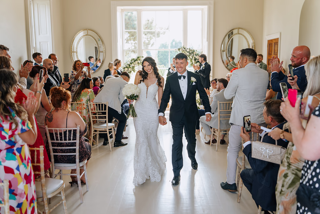 Bride holding a bouquet and groom in a black tuxedo with a bowtie walk back down the aisle after their ceremony at Stubton Hall