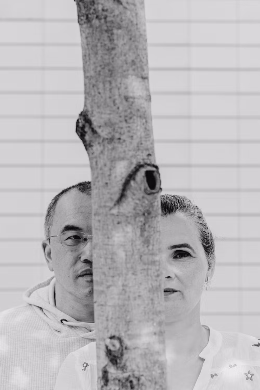 A greyscale photo of Martin & Daiga standing behind a tree, with the tree trunk vertically dividing their faces so each person is only half visible. The background is a white tiled wall.