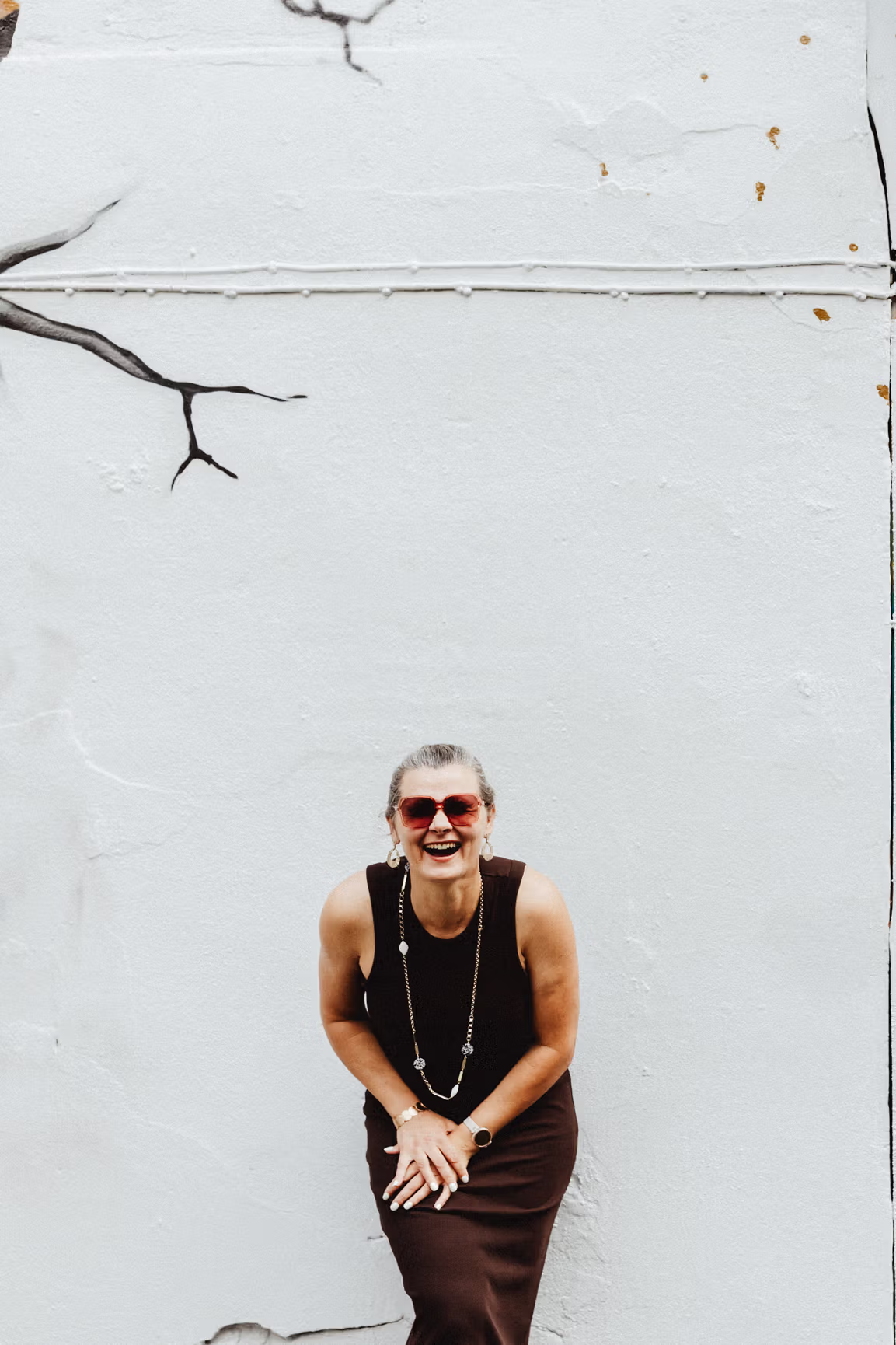 Daiga Cheung wearing a black dress and red sunglasses smiles and leans forward against a white wall with minimal black line art resembling a tree branch.