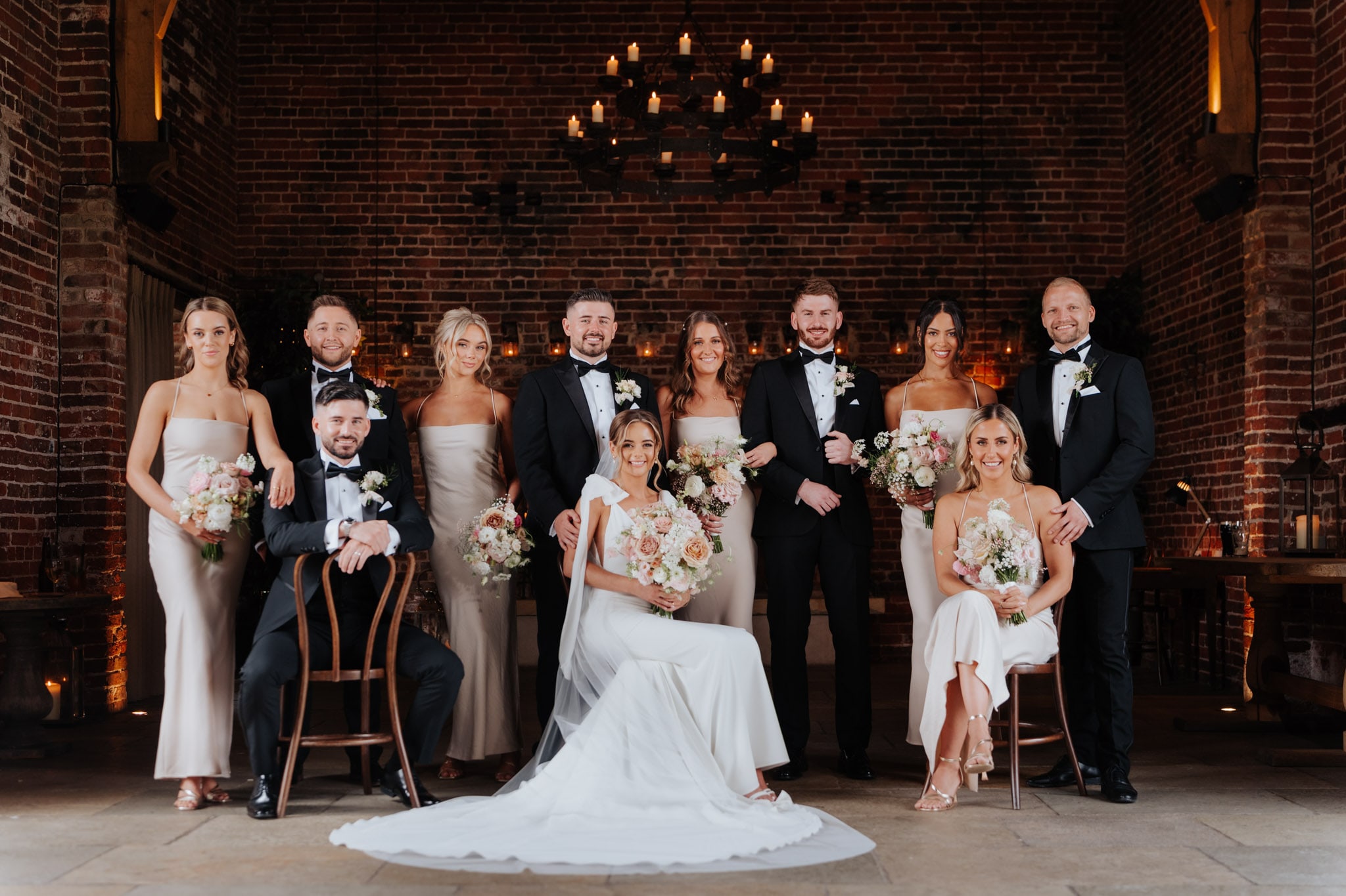 A wedding party poses indoors against a brick wall at Hazel Gap Barn. The bride sits in the center holding a bouquet, surrounded by smiling bridesmaids in light dresses and groomsmen in black tuxedos. Flowers and candles decorate the space.