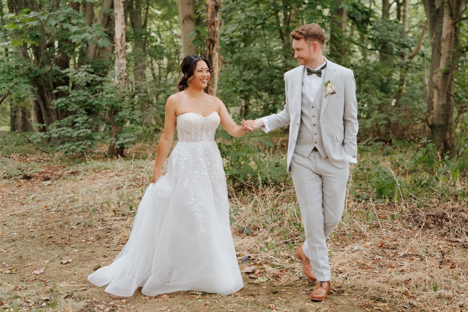 A bride in a strapless white gown and a groom in a light grey suit walk hand in hand in a wooded area, smiling at each other with trees and greenery in the background.