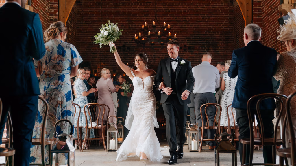 Bride walks down the aisle as she happiily points her bouquet into the air with groom in black tuxedo smiling next to her