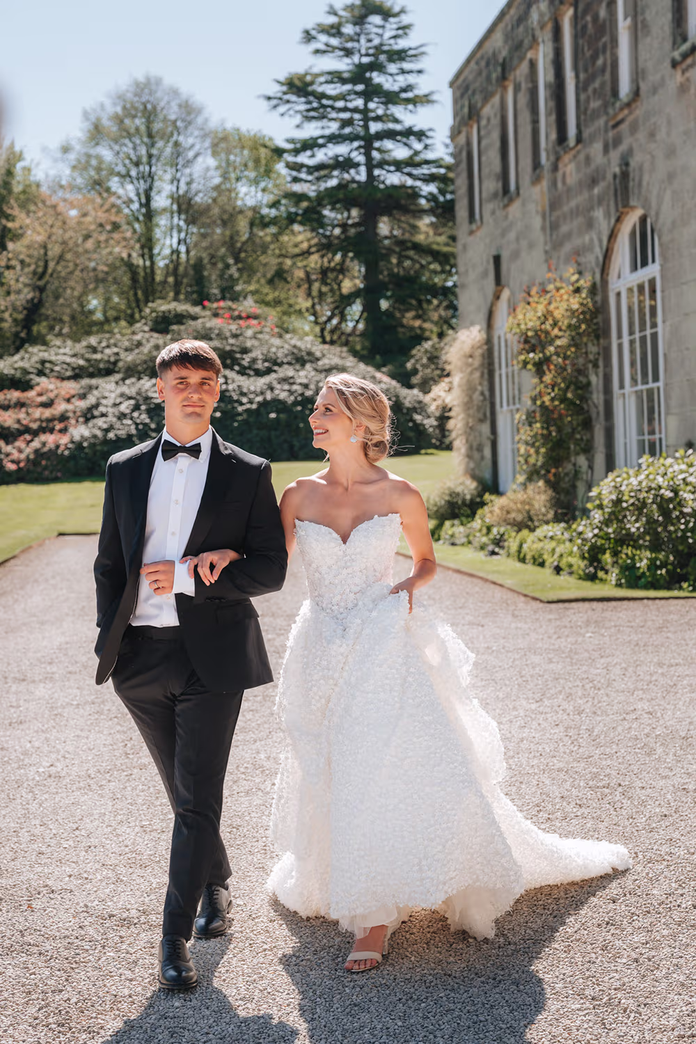 Bride in white dress & groom in black tuxedo walking arm in arm with a stately building behind them