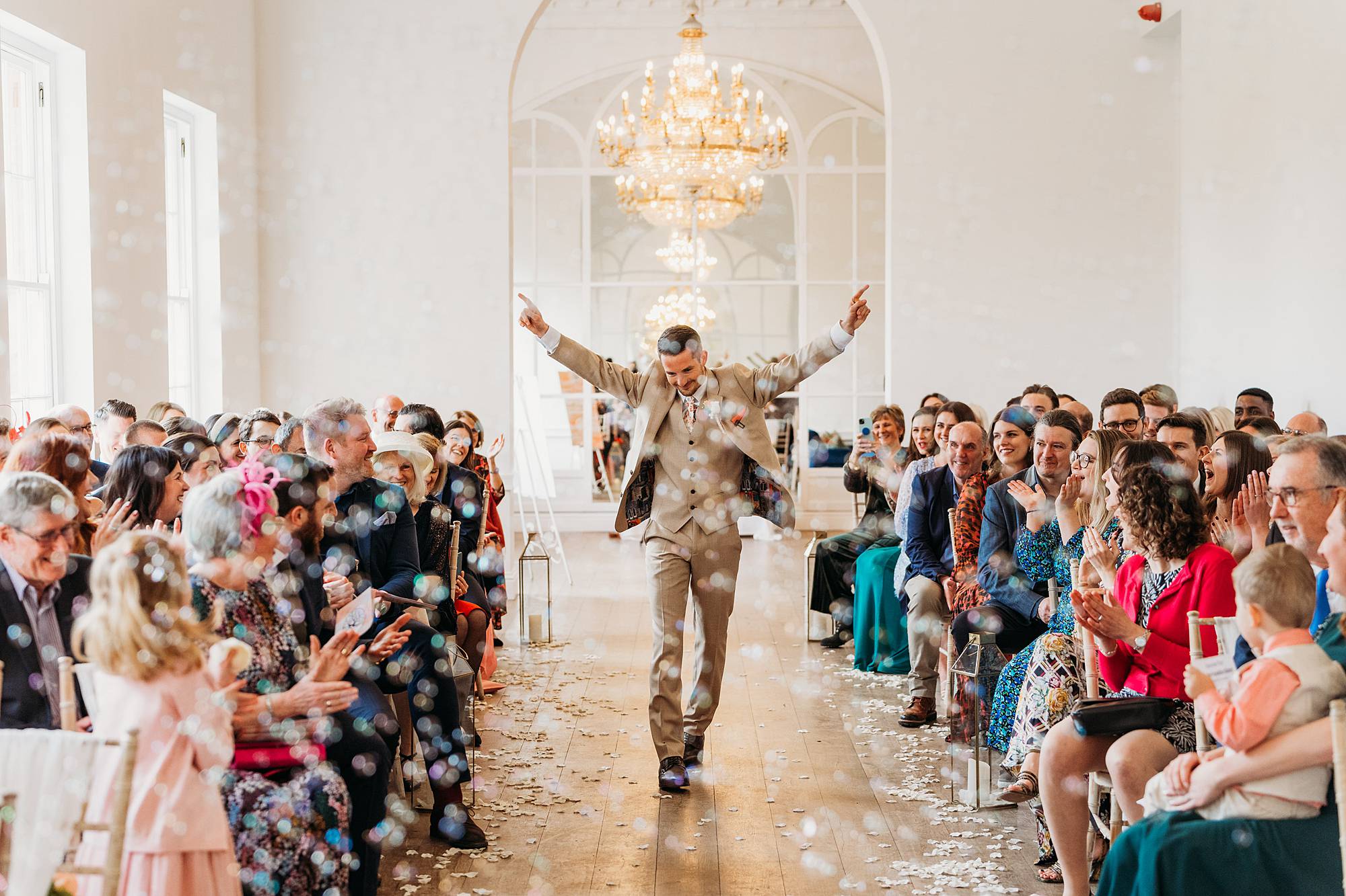 A groom in a beige suit walks joyfully down an aisle with arms raised, surrounded by smiling, applauding guests; bubbles and flower petals fill the air in a bright, elegant hall—captured perfectly by a Norwood Park Wedding Photographer.