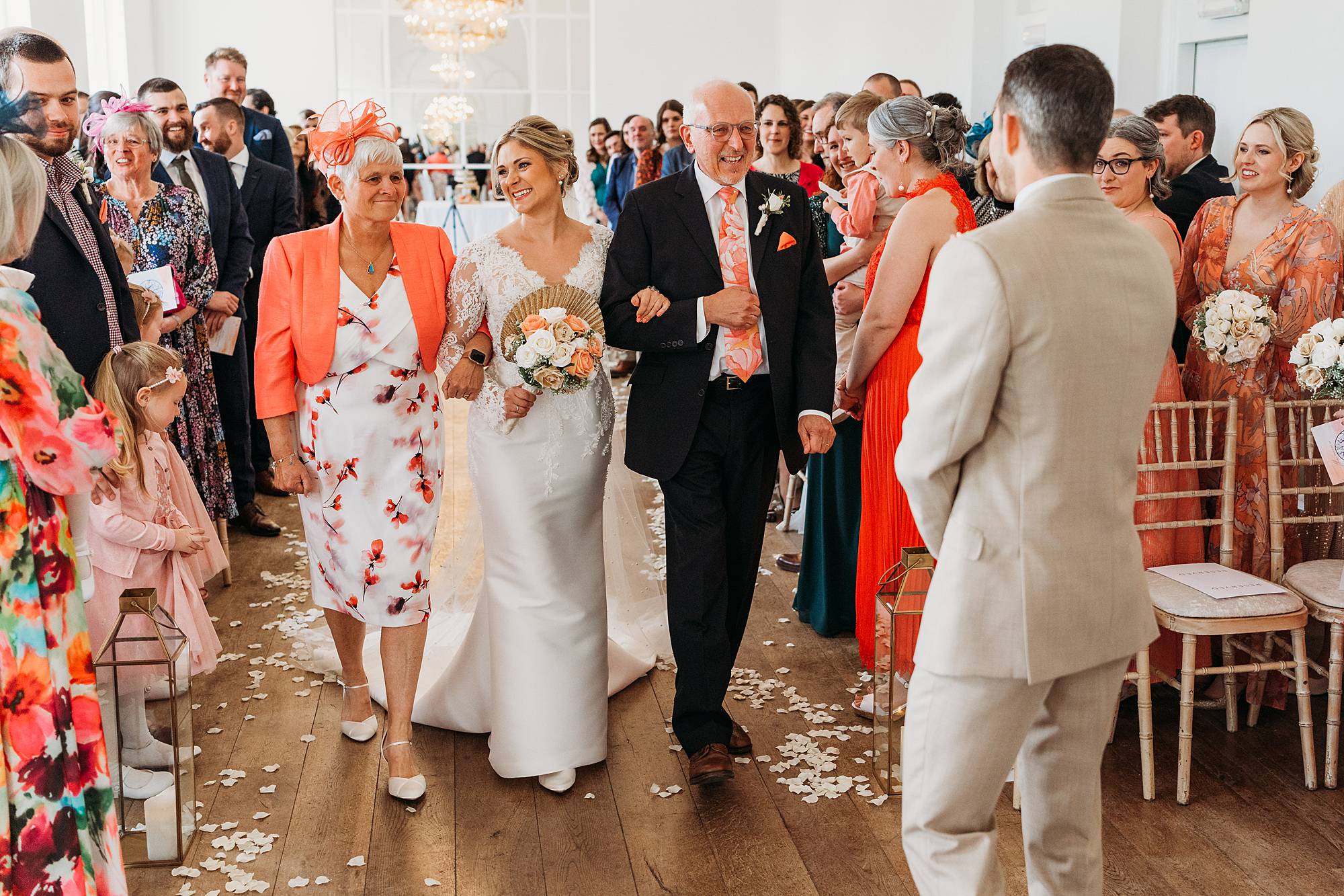 A bride in a white dress walks down the aisle with her parents at a Norwood Park wedding ceremony, surrounded by smiling guests in colourful attire and petals scattered on the floor.