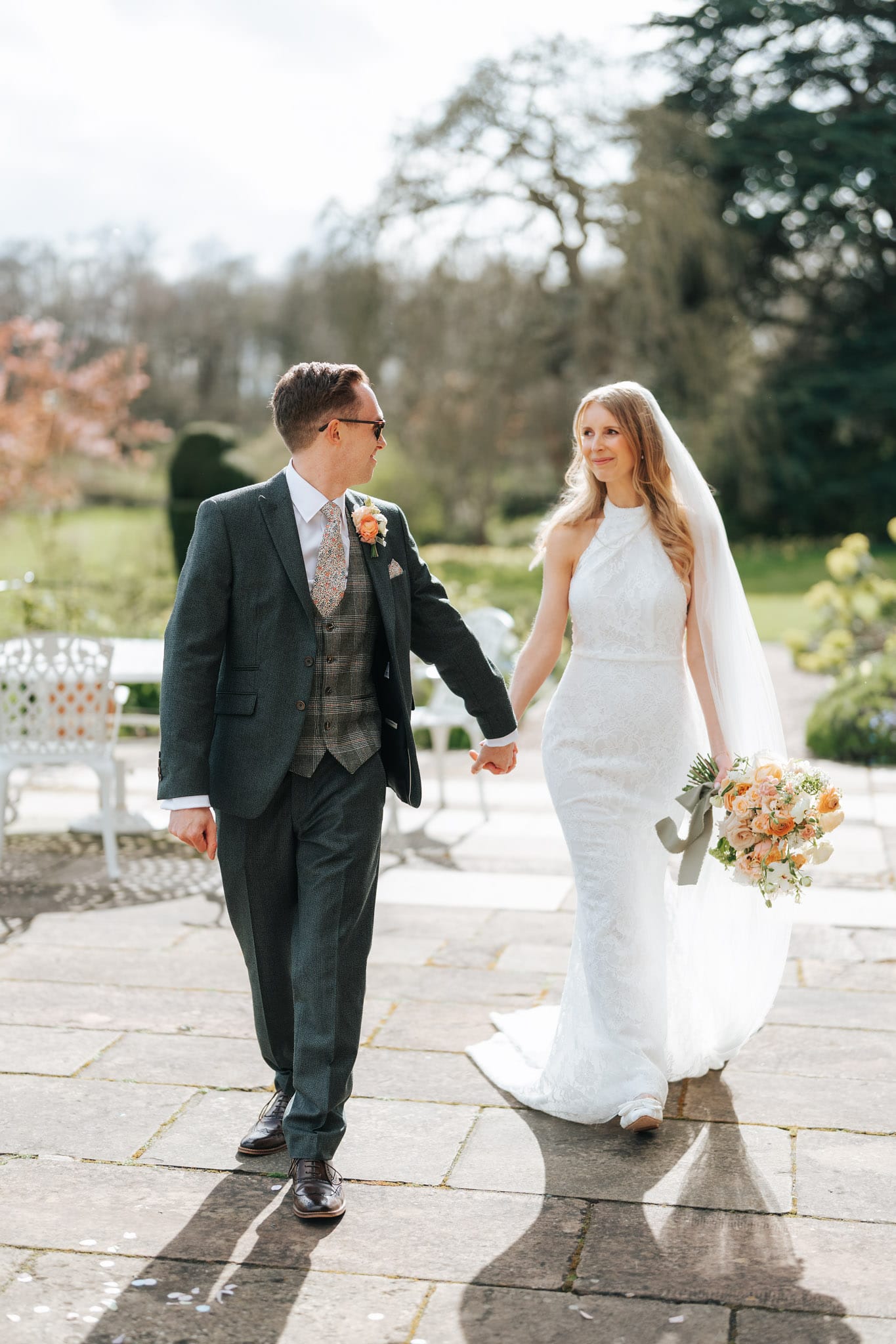 A bride in a white dress and veil holds a bouquet of flowers, walking hand in hand with her groom in a dark suit. Captured by a Norwood Park Wedding Photographer, they smile at each other outdoors on a sunny day surrounded by greenery.