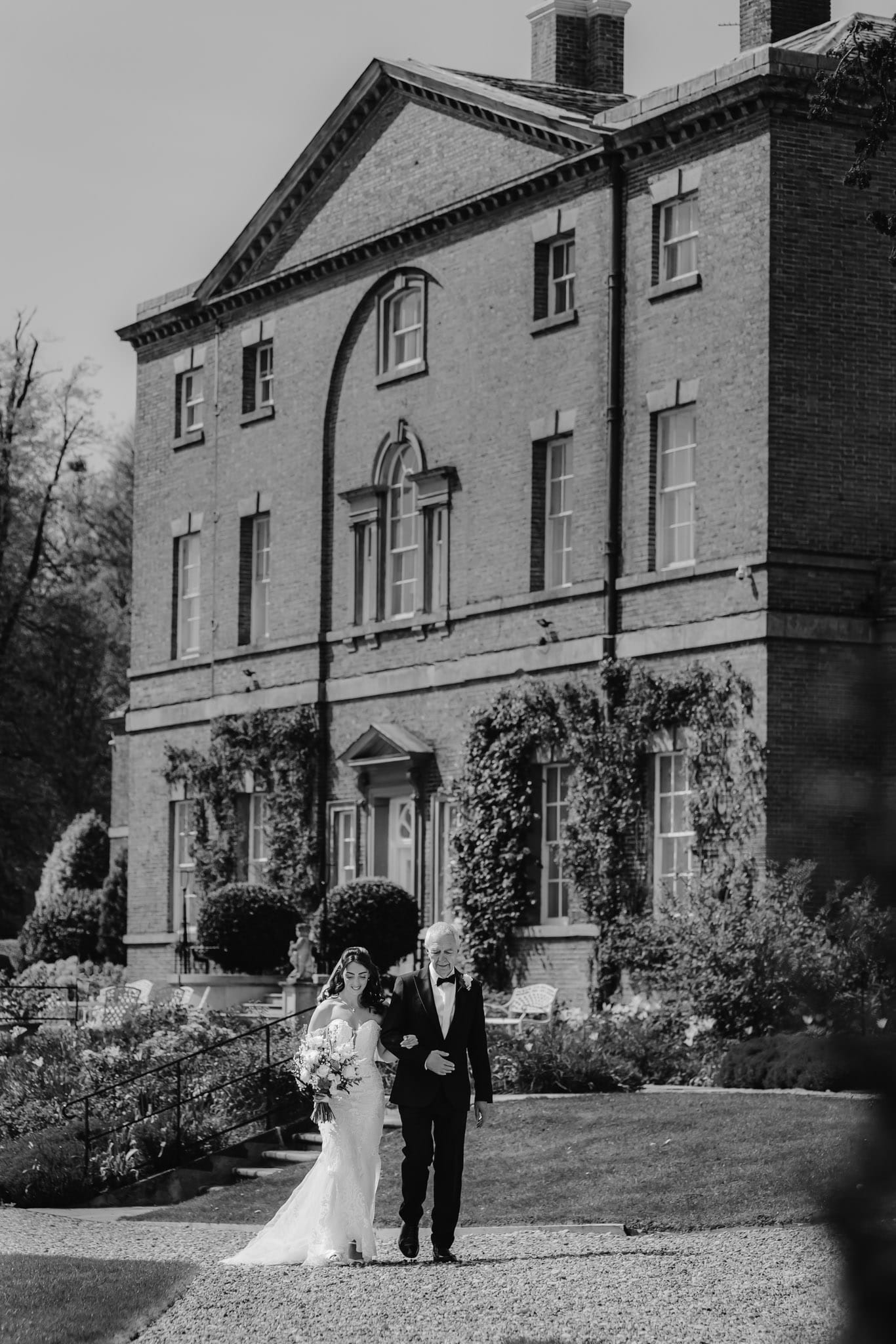 A bride walks arm in arm with her father to the wedding ceremony with Norwood Park in the background