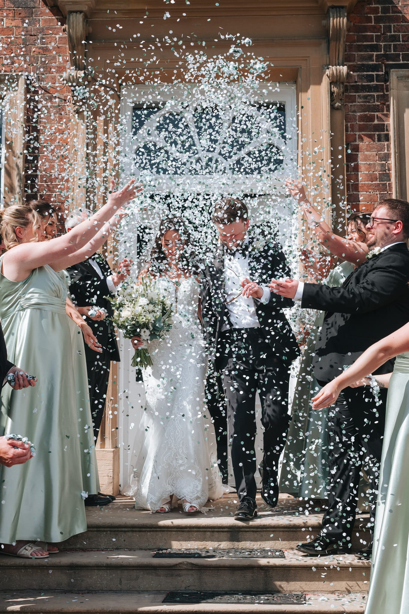 A bride and groom walk down steps outside Norwood Park as guests in formal attire throw confetti, celebrating their wedding. The Norwood Park Wedding Photographer captures the bride’s smile as she holds a bouquet amid a shower of white confetti.