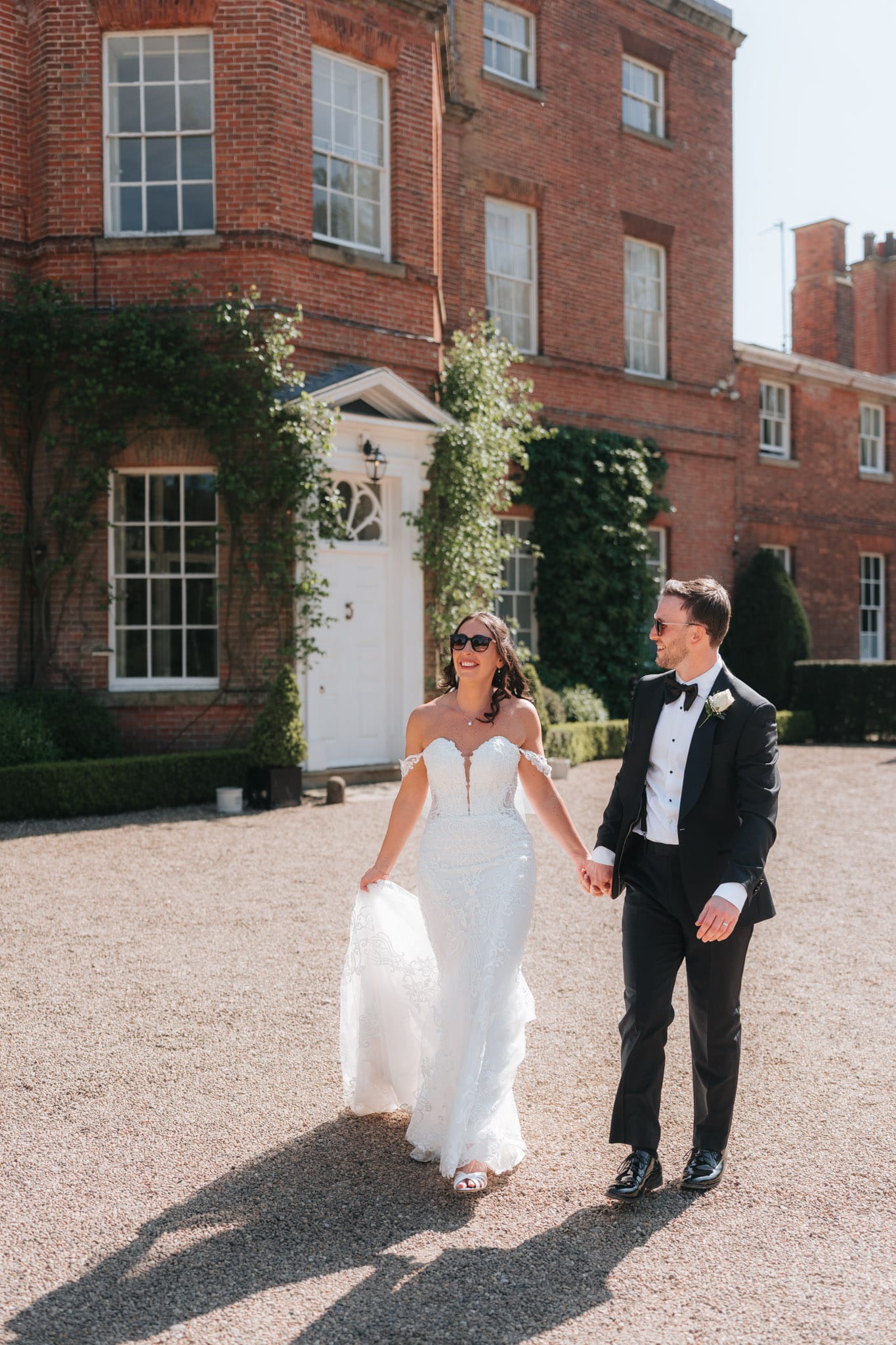 A bride in a white strapless gown and a groom in a black tuxedo walk hand in hand outside Norwood Park, smiling and enjoying a sunny day.