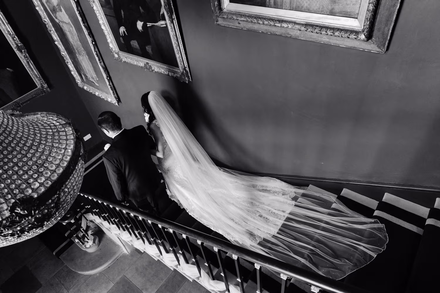 A bride in a long veil and a groom in a tuxedo walk down a staircase lined with framed portraits, captured by a Nottingham wedding photographer.